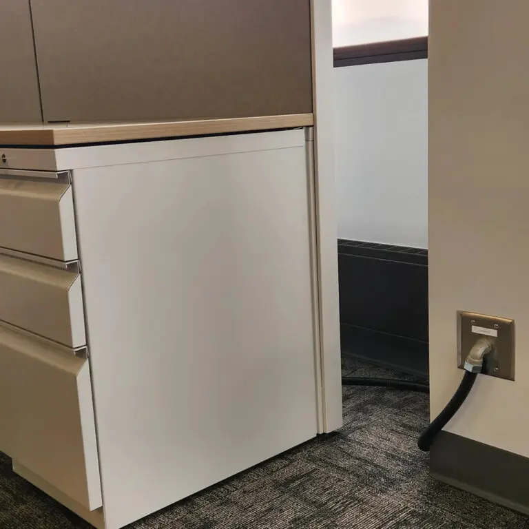 A white filing cabinet sits next to a wall with a power outlet and a thick black cable plugged in, likely set up by an electrician, on a gray carpeted office floor.