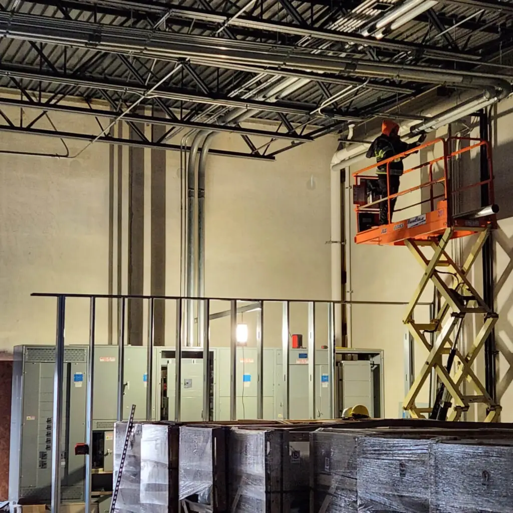 An electrician in a hard hat stands on an elevated scissor lift, performing maintenance near the ceiling of an industrial facility with electrical panels and pipes below.