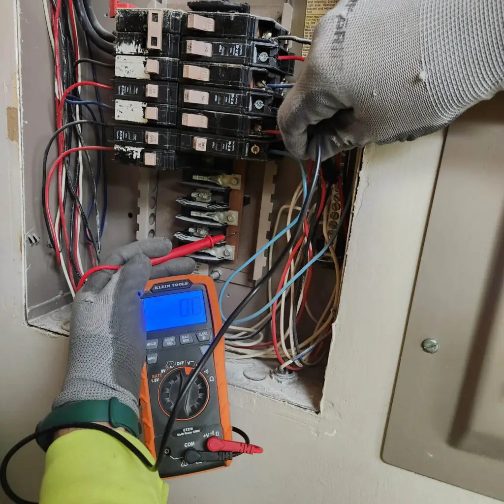 A gloved electrician uses a digital multimeter to measure voltage in an electrical panel with exposed wires and circuit breakers.