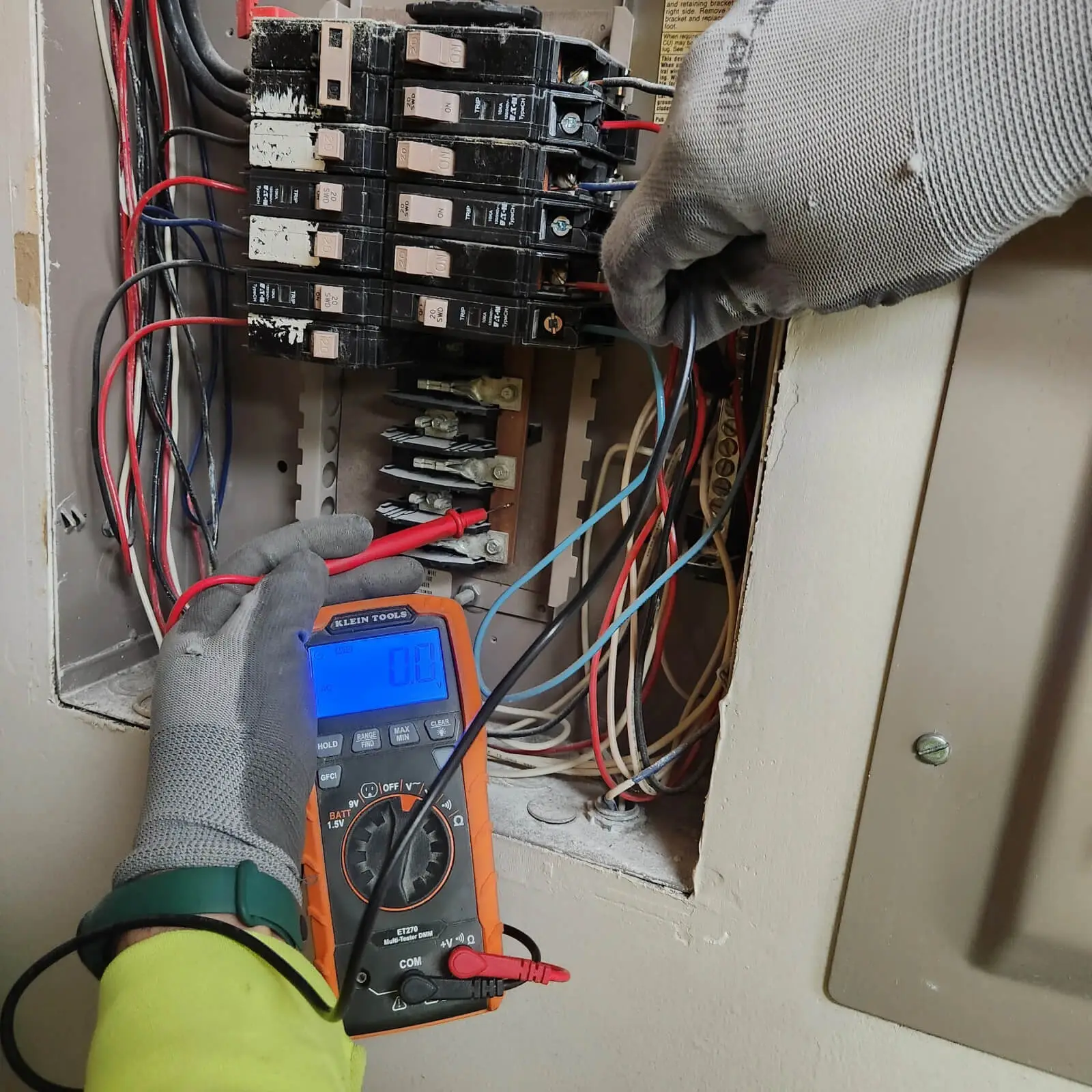 A gloved electrician uses a digital multimeter to measure voltage in an electrical panel with exposed wires and circuit breakers.