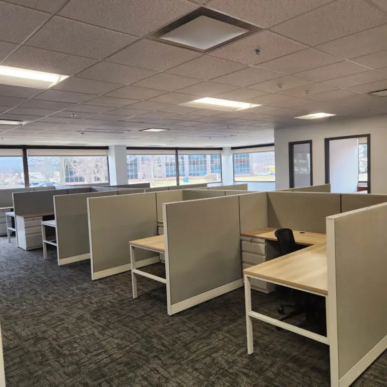 An empty modern office space with several cubicles, desks, and chairs, large windows letting in natural light, and neutral-colored walls and carpet—ready for an electrician to install customized lighting or power solutions as needed.