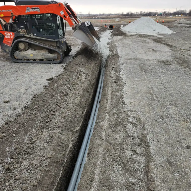 An orange skid steer loader dumps gravel into a trench containing gray pipes at a construction site, where an electrician might later install wiring; a pile of gravel and open land can be seen in the background.