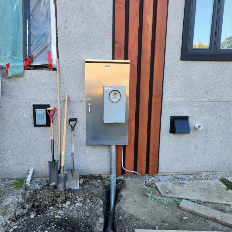 A house wall with an electric meter, three shovels leaning nearby, exposed wiring—likely awaiting work by an electrician—and a trench in the ground below the meter. Construction materials are visible around the site.