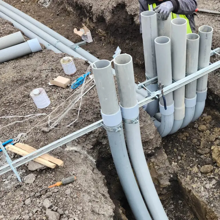 Multiple gray PVC conduit pipes are grouped and arranged in trenches for underground electrical installation, with tools and materials scattered nearby on the soil. An electrician’s gloved hands are partially visible at the top.
