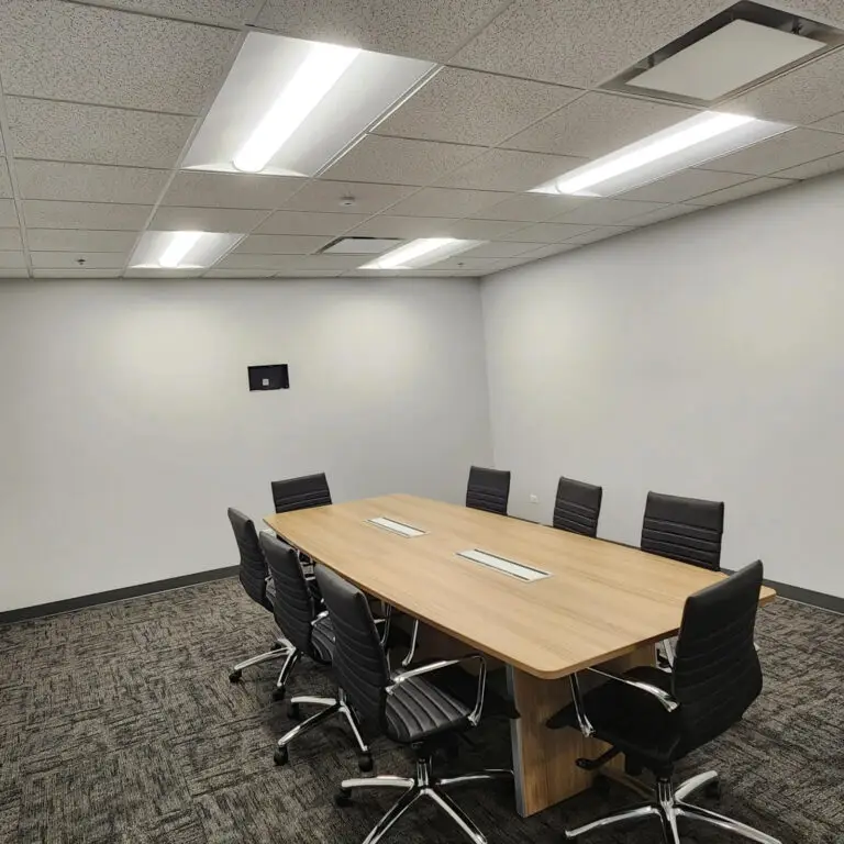A modern conference room with a wooden table, eight black swivel chairs, gray carpet, white walls, and bright overhead lighting installed by a skilled electrician. A small screen is mounted on one wall.