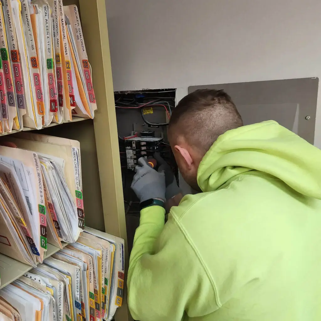An electrician in a yellow hoodie and gloves works on an open electrical panel near shelves filled with labeled file folders.