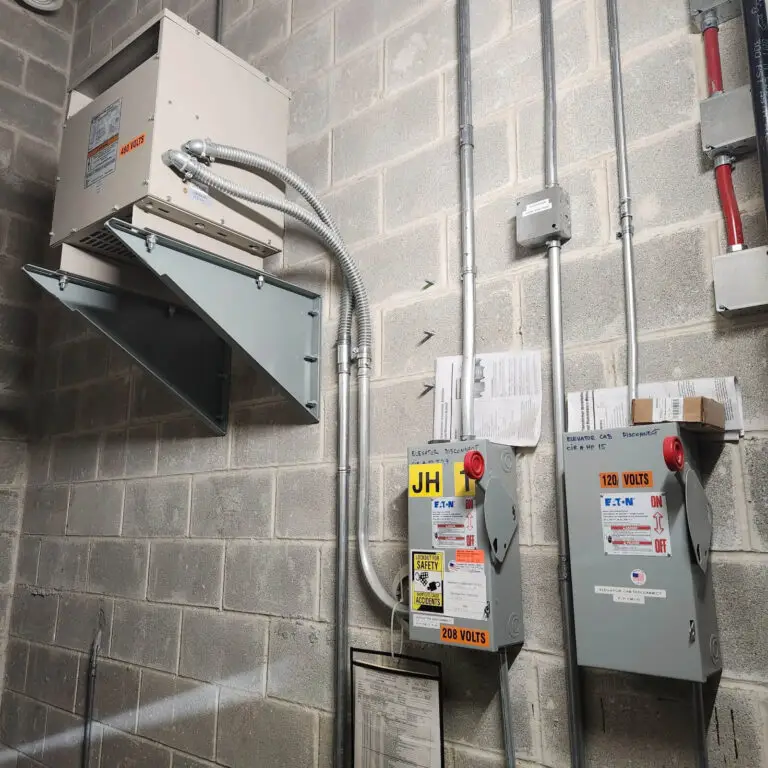 Two electrical panels labeled 120 volts and 208 volts are mounted on a concrete wall, with warning stickers added by the electrician. Above them is an air handling unit with metal ducts and conduit piping attached. Papers are posted on the wall nearby.