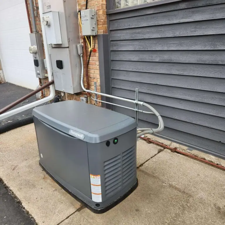 A gray standby generator is installed next to a brick wall and gray garage door, with electrical panels and cables mounted above it on the wall.