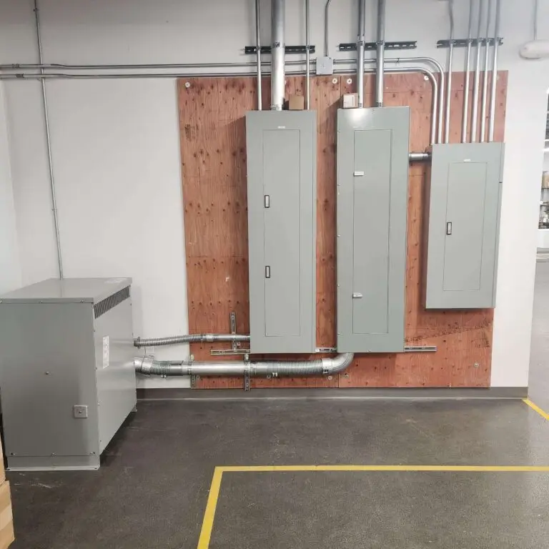 Three gray electrical panels mounted on a wooden board with metal conduits, next to a large gray electrical box on a black floor marked with yellow lines.