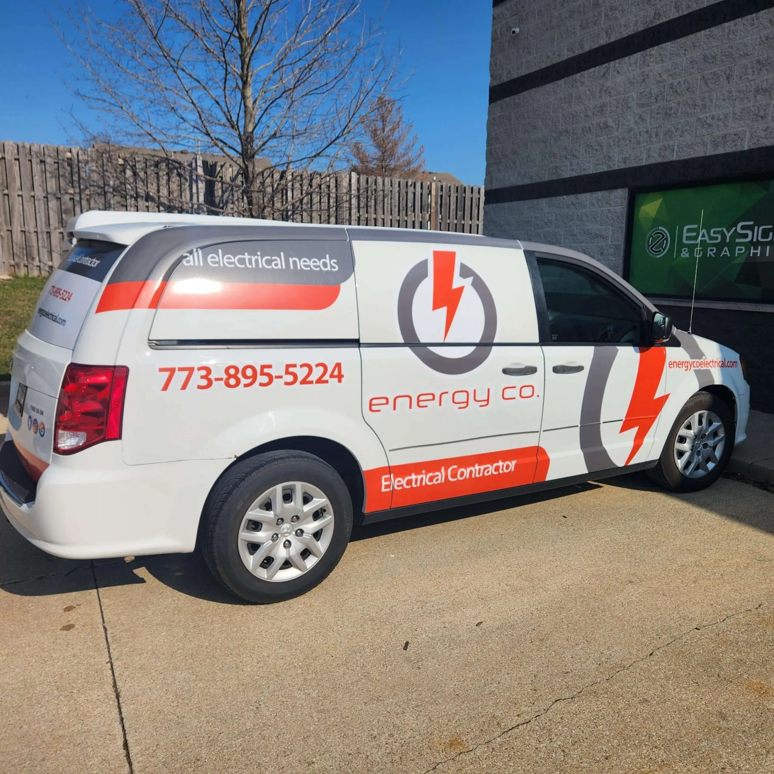A white van with red and gray graphics for energy co. Electrical Contractor, parked by a building. The van displays a phone number, lightning bolt logo, and all electrical needs text.