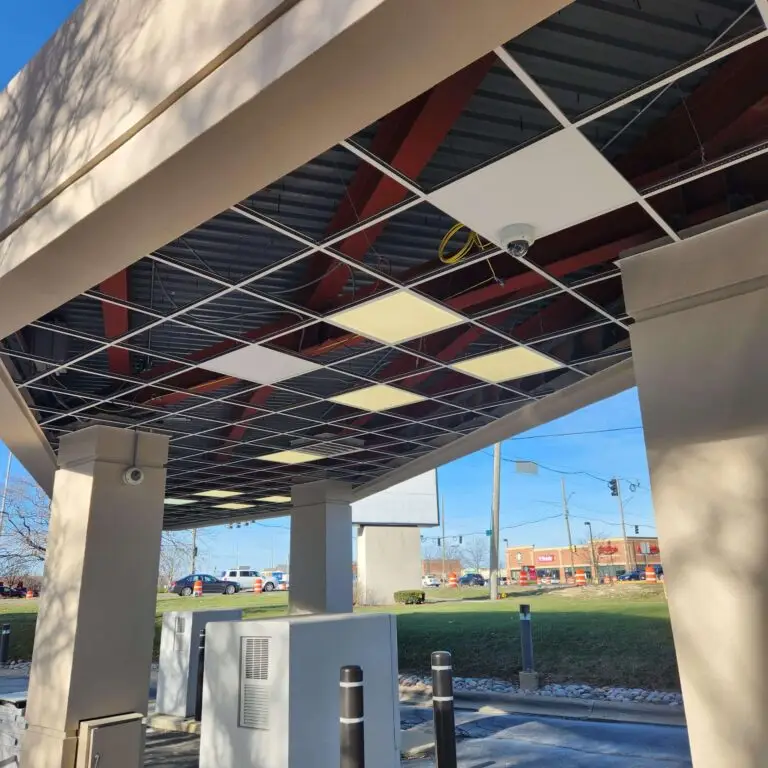 A view from beneath a drive-thru canopy with missing ceiling tiles, exposing red beams and wiring. In the background, a street, cars, and buildings are visible on a sunny day.