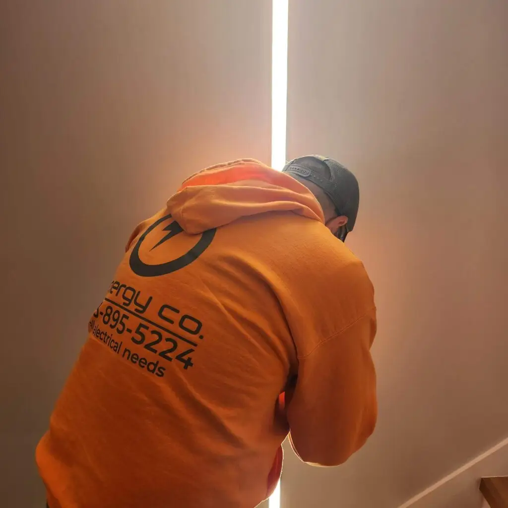 A person in an orange hoodie with a company logo and phone number is working near a bright vertical light strip on a wall.