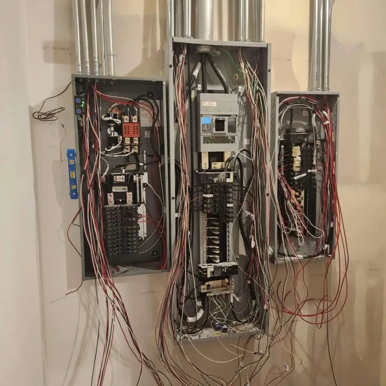 Three open electrical panels filled with circuit breakers and many exposed wires hanging and running between them, located indoors against a beige wall.