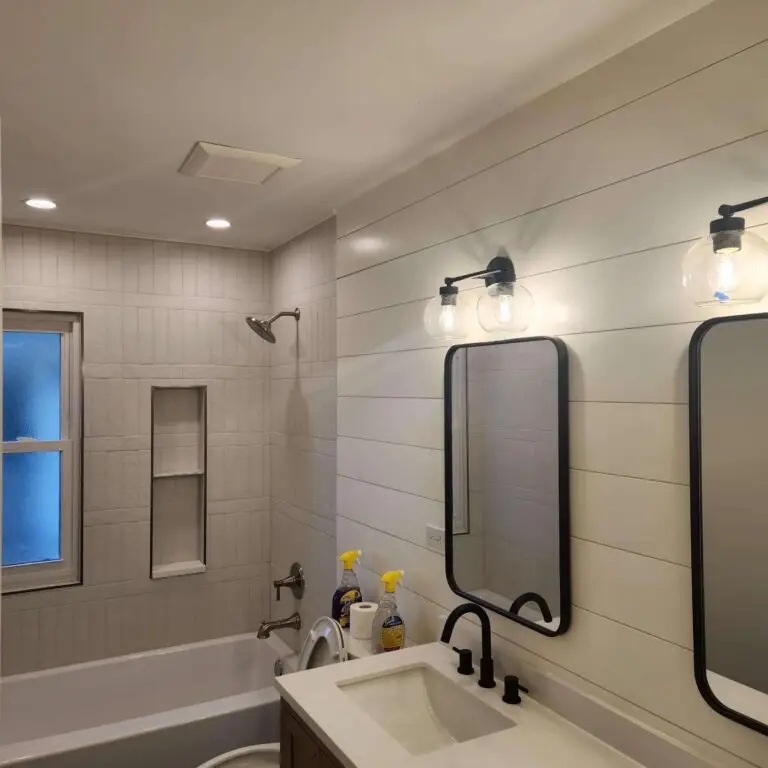Modern bathroom with shiplap walls, two mirrors above a sink, black fixtures, and a bathtub with a tiled shower. Cleaning supplies are on the sink. Ceiling lights and frosted window provide illumination.