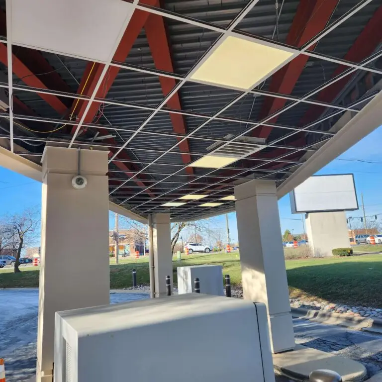 A bank drive-thru area with exposed ceiling tiles and wires, supported by columns. Some ceiling panels are missing or out of place. Outdoor bank equipment is visible, with cars and a grassy area in the background.