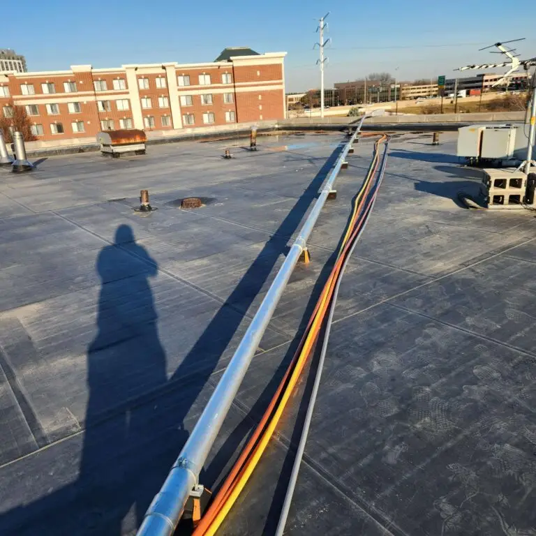 A rooftop with several colored cables running through metal conduit, casting a long human shadow. In the background, there is a red brick building, power lines, and clear blue sky.