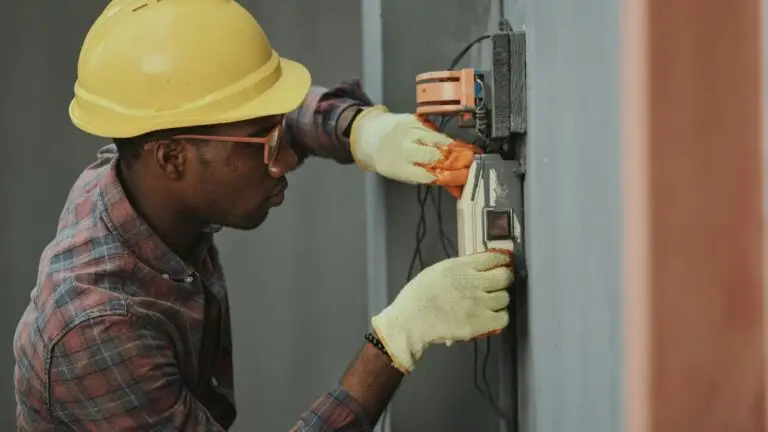 An industrial electrician in Chicago, wearing a yellow hard hat and gloves, works on an electrical panel attached to a gray wall.