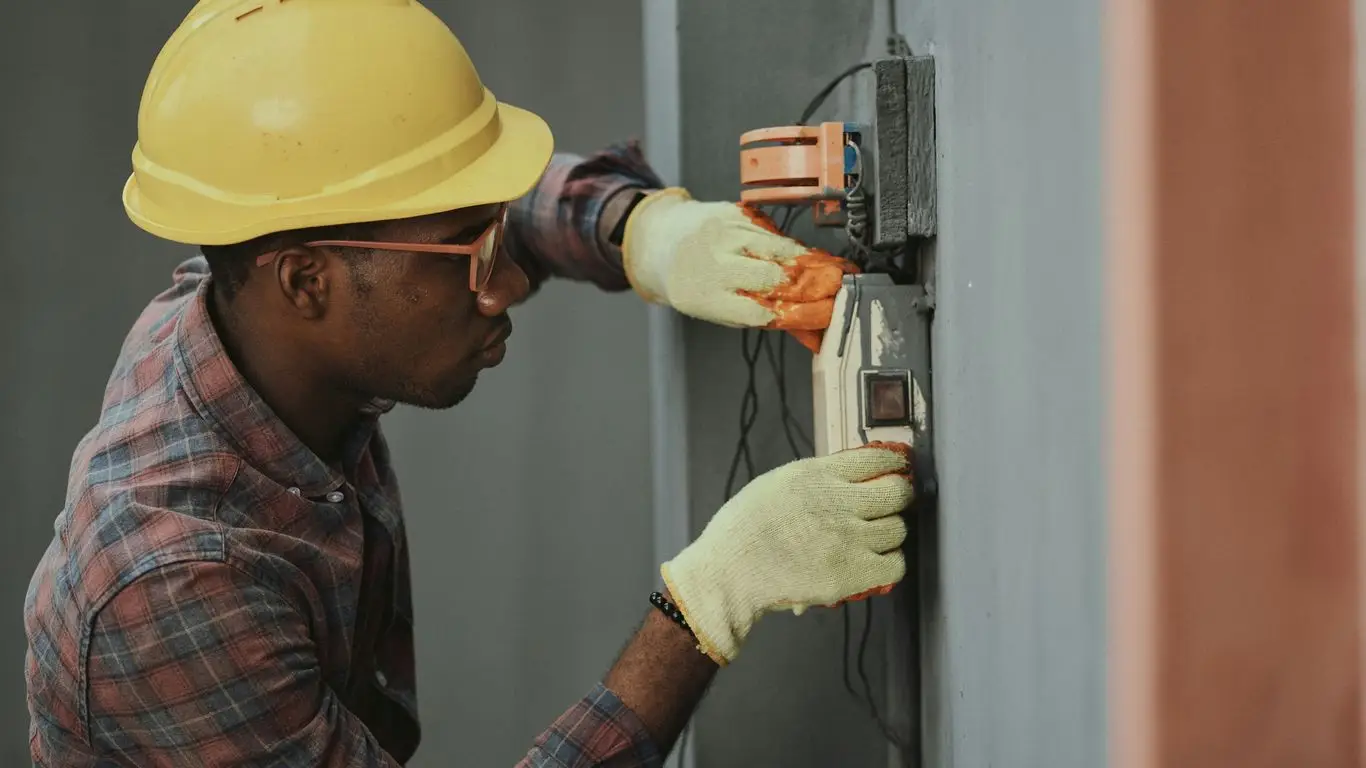 An industrial electrician in Chicago, wearing a yellow hard hat and gloves, works on an electrical panel attached to a gray wall.