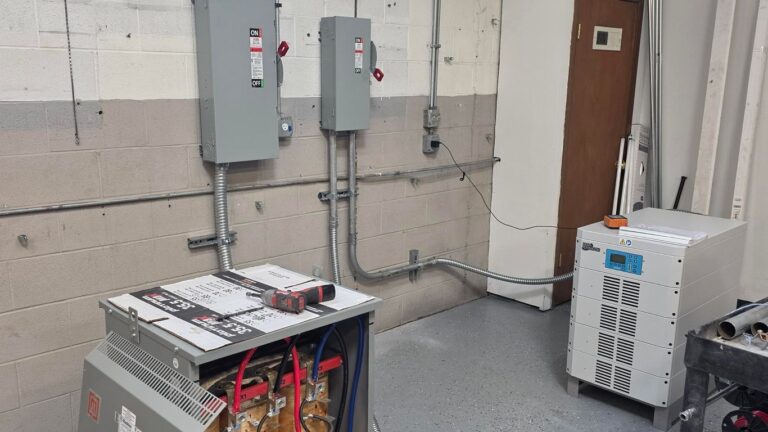 Industrial electrical room with wall-mounted panels, exposed conduit, a transformer with tools on top, and a white equipment unit with a paper on it, all set on a gray concrete floor near a wooden door—an ideal setting for comprehensive electrical installation service in Chicago.