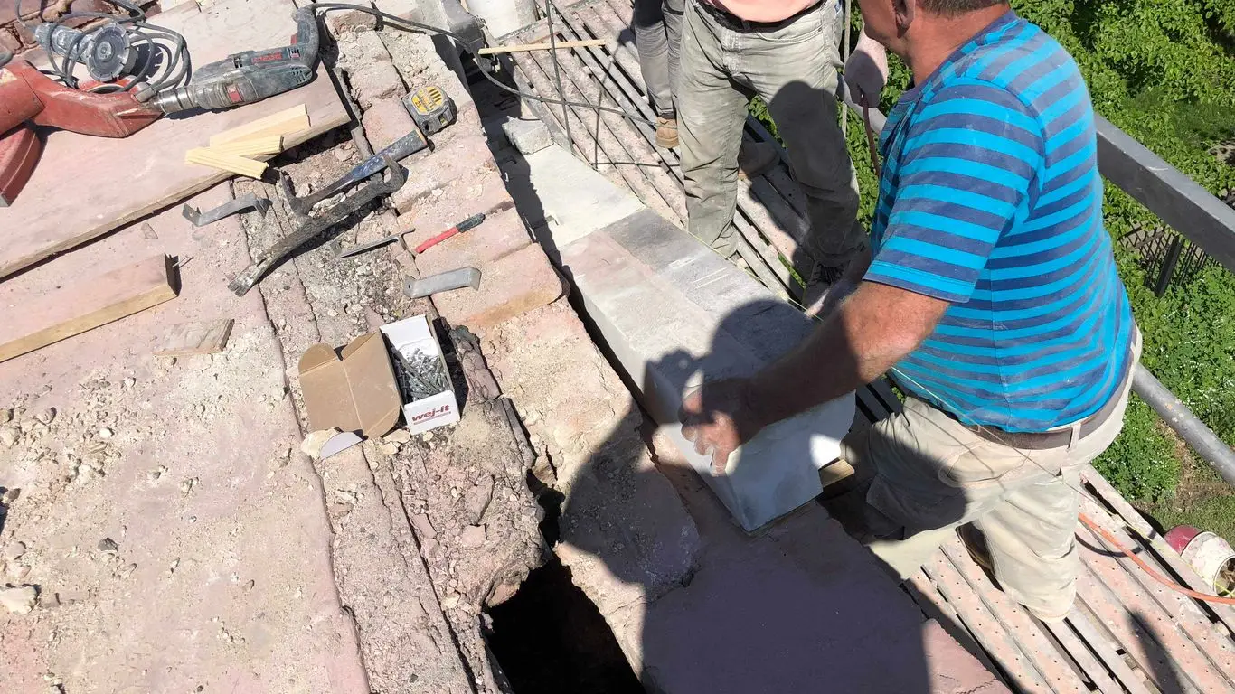 Two workers in casual clothes, reminiscent of a skilled electrician Chicago might employ, repair a damaged concrete surface on scaffolding, surrounded by tools, a power drill, and a box of screws, with greenery visible to the right.