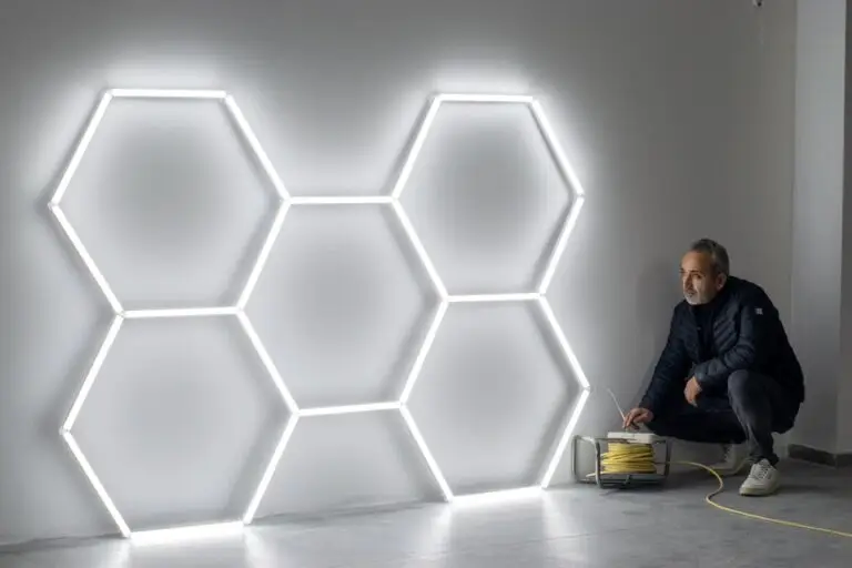 A person crouches near a wall with six hexagons made of glowing white LED tubes. The person is plugging in a yellow extension cord connected to the illuminated artwork, which showcases the innovation possible through commercial lighting retrofit in Chicago.