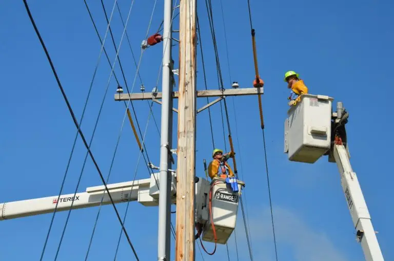 Two utility workers in safety gear, employed by commercial electric companies, work from cherry picker buckets on a power pole, surrounded by electrical wires, under a clear blue sky.