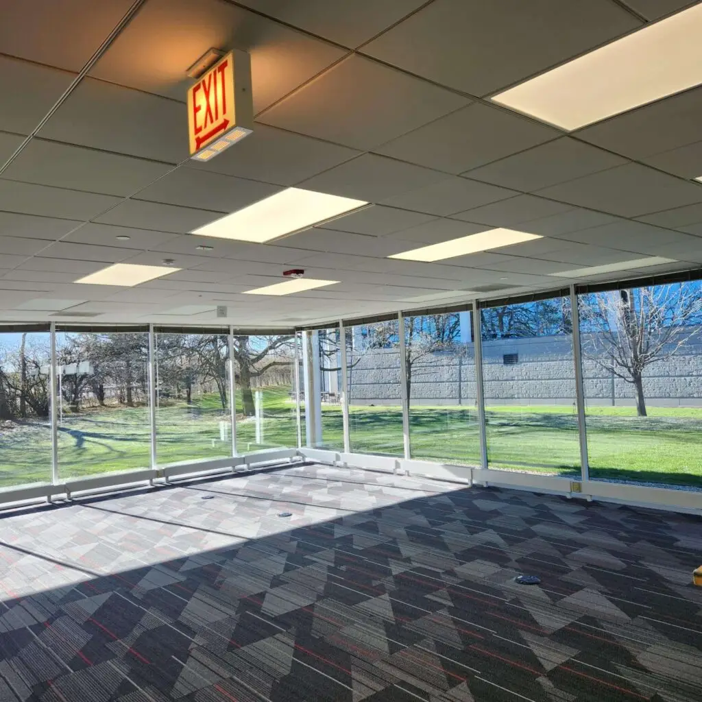 A sunlit, empty room with large windows, carpeted floor, and an exit sign on the ceiling. Trees and grass are visible outside through the glass.
