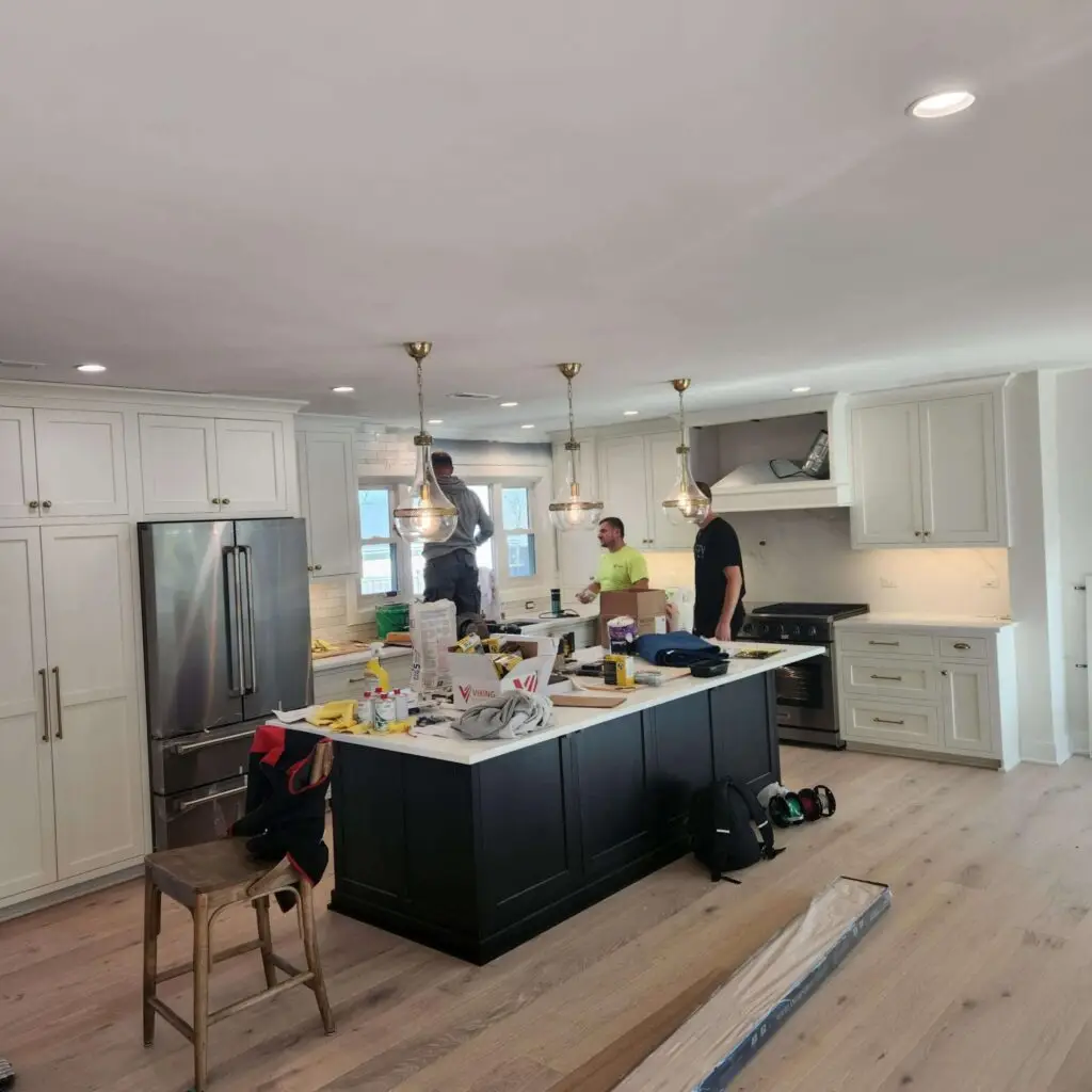 Three people work in a modern kitchen with white cabinets, a black island, and hanging lights. Tools and supplies are scattered on the island and floor, suggesting ongoing renovation or installation work.