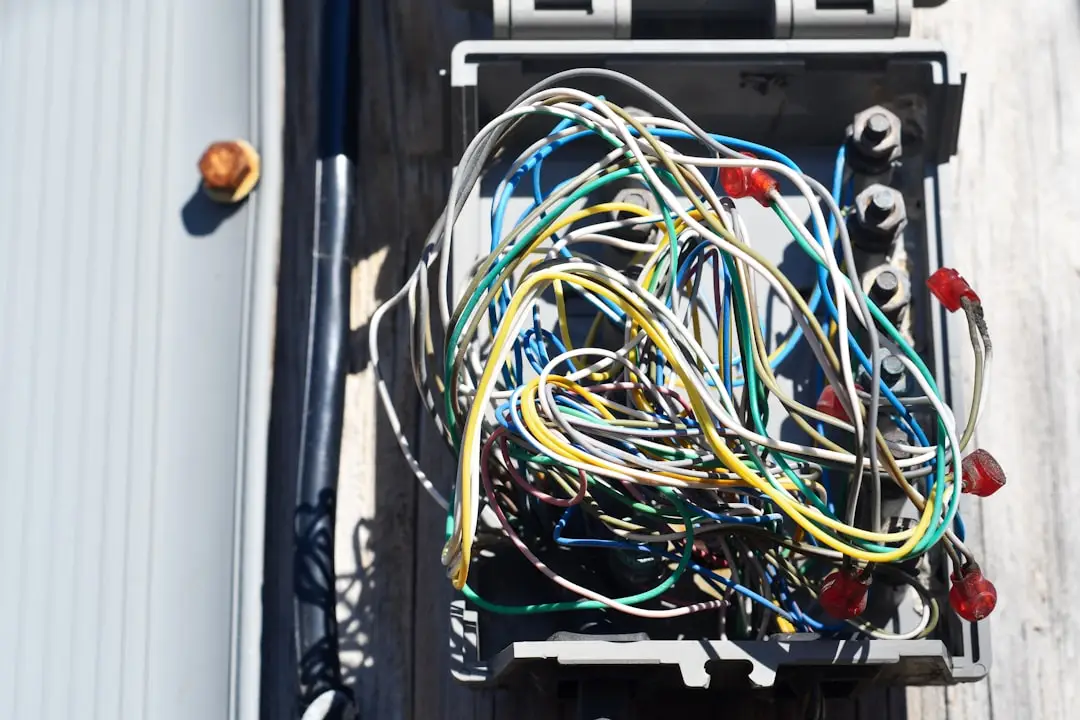 An open electrical box filled with tangled, multicolored wires and connectors—including a conspicuous hot tub electrical cable—mounted on a wooden surface in bright sunlight.