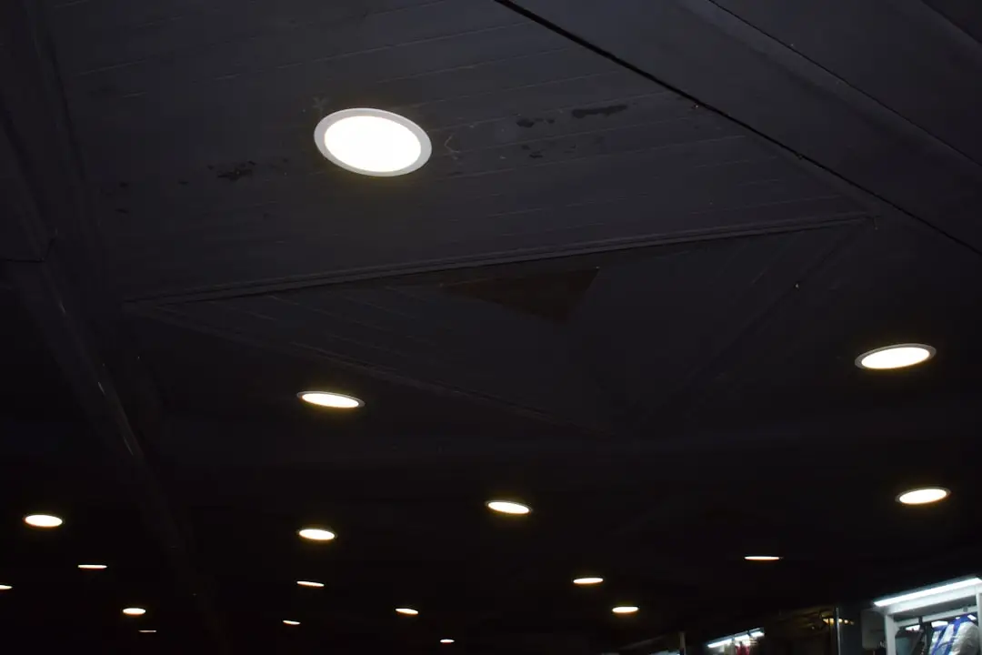 A dark ceiling with several circular LED recessed lighting retrofit fixtures glowing. The lights are spaced out, and parts of the ceiling have geometric panel designs. Some reflections and light fixtures are visible in the lower right corner.