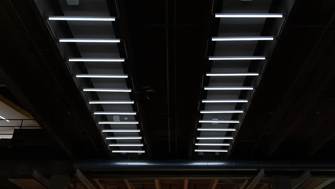 Two parallel rows of evenly spaced, horizontal retrofit fluorescent lighting LED fixtures are mounted on a dark industrial ceiling, creating a symmetrical pattern.