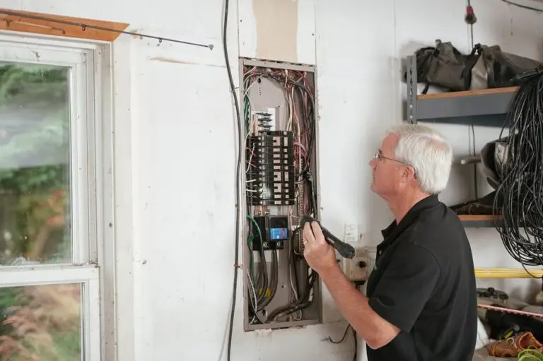 An older man with gray hair inspects an open electrical panel in a garage, holding a flashlight and wearing glasses. Exposed wires are visible, highlighting the importance of finding a licensed electrician near me for safe repairs. A window and shelves with tools are nearby.