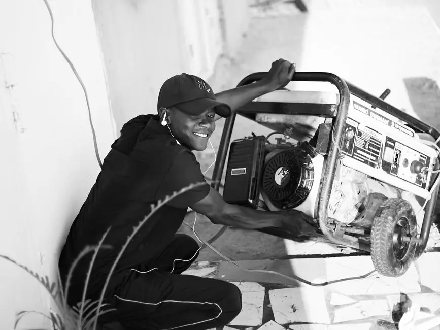 A person wearing a cap and earphones smiles while holding and adjusting a portable generator outdoors, with sunlight casting shadows on the ground, perhaps preparing for a generator repair service.