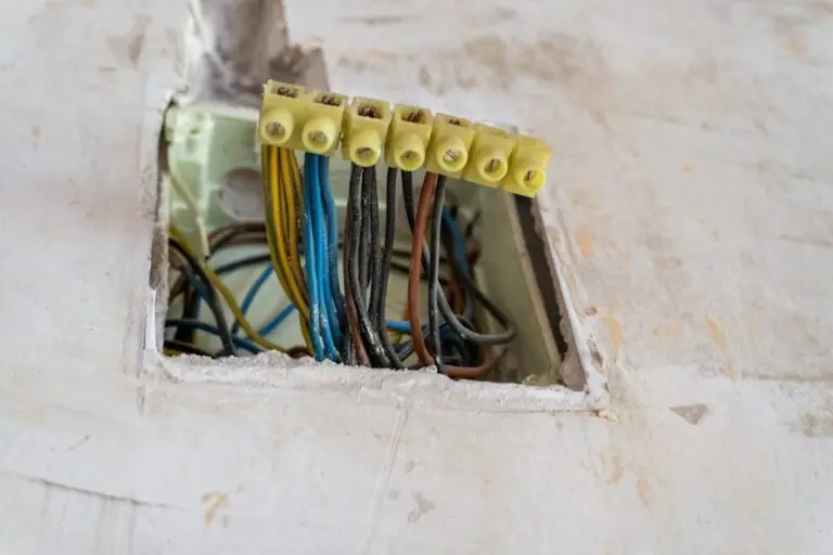 An open square cutout in a chipped white plaster wall exposing an old residential electrical junction box, with multiple color-coded wires — blue, brown, yellow, and black — twisted together and capped with yellow wire nuts, illustrating how residential electrical wiring runs from the meter box to the receptacle behind the wall.