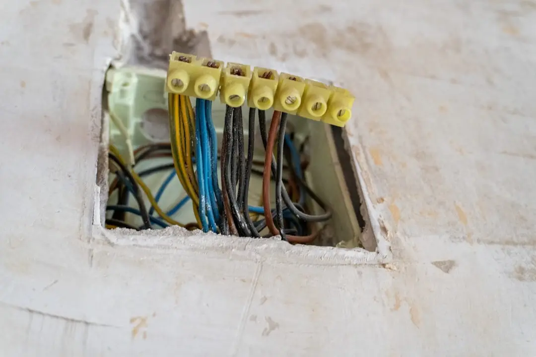 An open square cutout in a chipped white plaster wall exposing an old residential electrical junction box, with multiple color-coded wires — blue, brown, yellow, and black — twisted together and capped with yellow wire nuts, illustrating how residential electrical wiring runs from the meter box to the receptacle behind the wall.