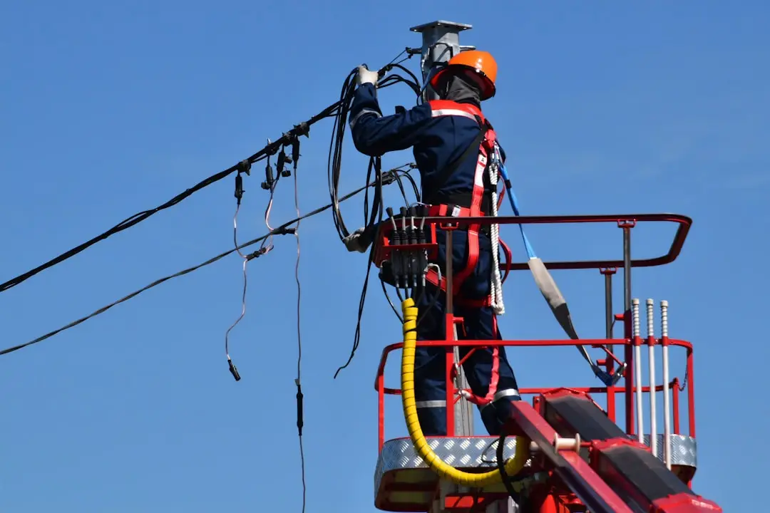 A commercial electrician in a dark blue high-visibility jumpsuit and orange hard hat, secured with a safety harness, working from a red aerial bucket lift to repair a tangle of black overhead power cables against a clear blue sky, showing the kind of high-voltage line repair a licensed commercial electrician near me handles on emergency call-outs.